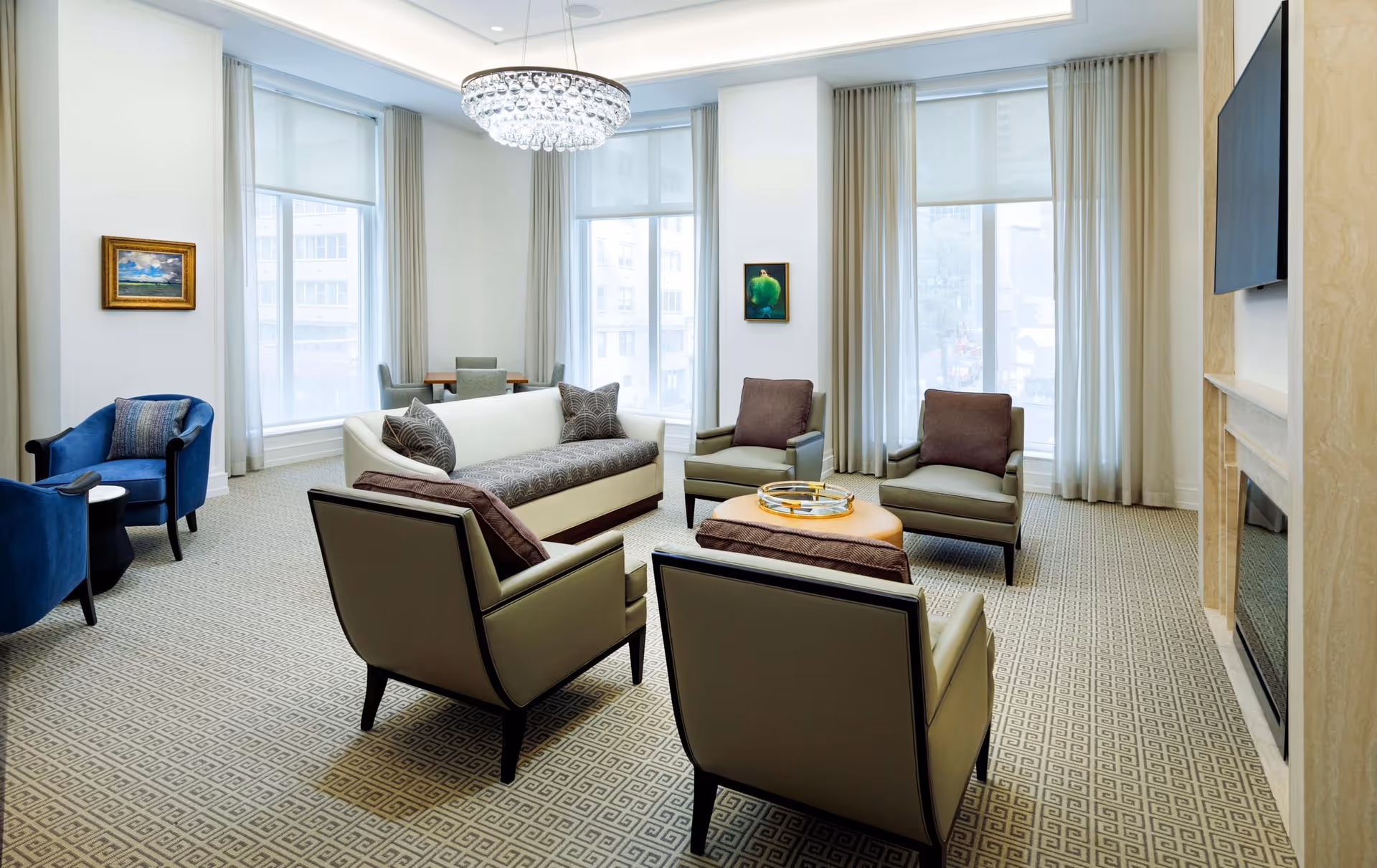 A bright and elegant living room with large windows covered by sheer curtains, a crystal chandelier, a white sofa with patterned cushions, four armchairs arranged around a round ottoman, a fireplace with a mounted flat-screen TV, and two framed paintings on the walls.