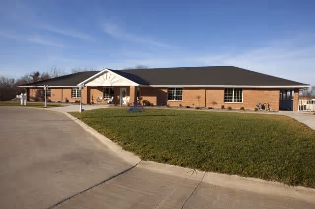 Single-story brick building with a covered entrance, surrounded by a paved driveway and a grassy area under a clear blue sky.