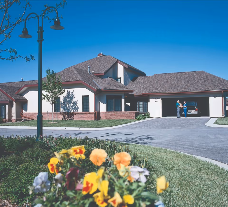Exterior view of a single-story building with a sloped roof, a driveway leading to a garage where two people are standing and talking. In the foreground, there is a garden with colorful flowers and a street lamp. The sky is clear and blue.