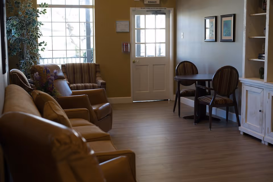 A cozy interior room with a row of brown leather and fabric chairs along the left side, a small round table with two striped chairs on the right, a white door with glass panels at the back, and a large window letting in natural light. There are framed pictures on the wall and a white cabinet with shelves on the right side.