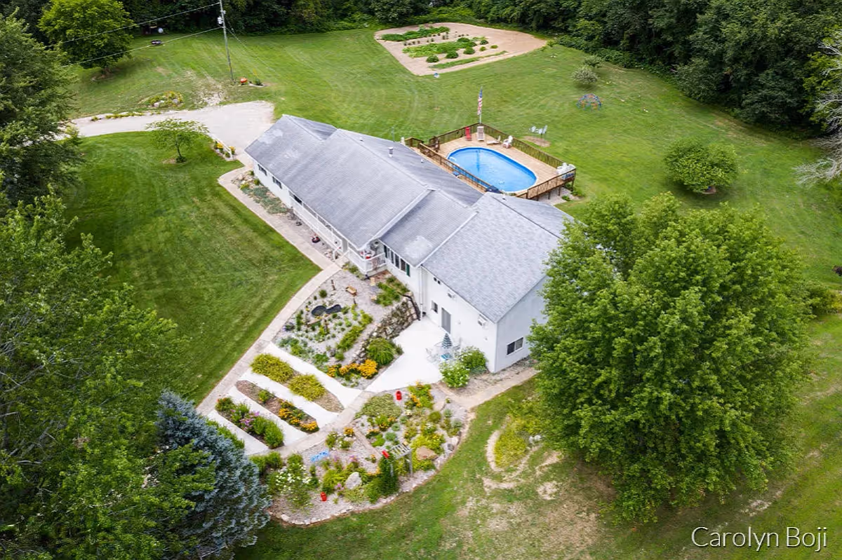 Aerial view of a single-story building surrounded by well-maintained green lawns and gardens. The building has a gray roof and a small outdoor swimming pool with a wooden deck. There are various trees and landscaped flower beds around the property.