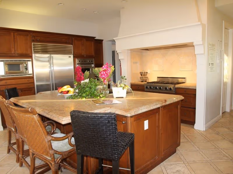 A spacious kitchen with a large island countertop surrounded by four chairs. The kitchen features wooden cabinets, a stainless steel refrigerator, a built-in microwave, and a stove with a decorative hood. There are plants and flowers placed on the island, and the floor is tiled.