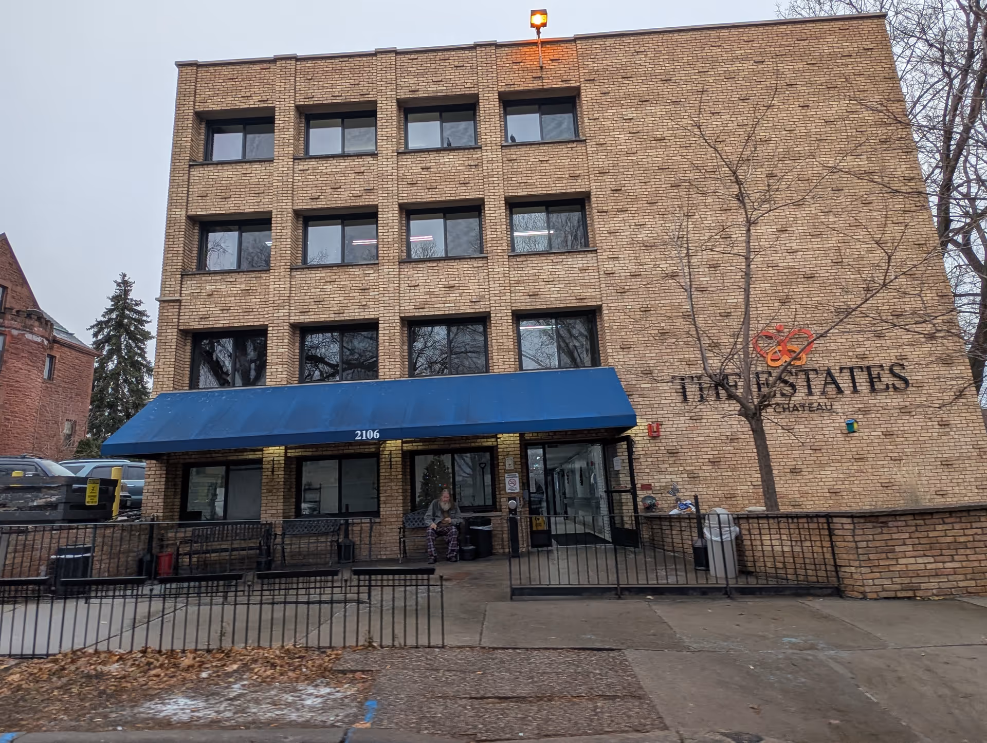 Exterior view of a three-story brick building with a blue awning over the entrance. The building has multiple windows and a sign on the right side that reads 'The Estates at Chateau'. A person is sitting on a bench near the entrance. Leafless trees and parked cars are visible around the building.