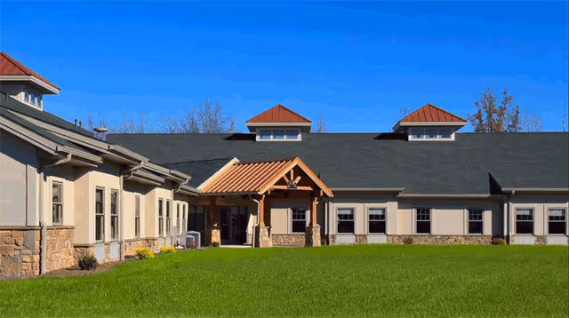 Exterior view of a single-story building with beige walls, stone accents, and a dark shingled roof with two small cupolas. The entrance features a wooden gabled porch. The building is surrounded by a well-maintained green lawn under a clear blue sky.