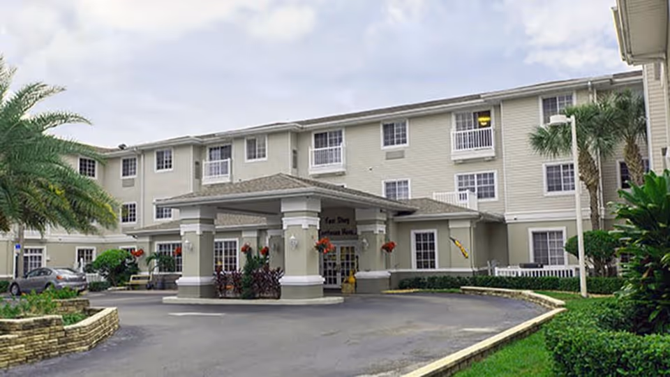Exterior view of Sonata Vero Beach, a three-story senior living facility with a covered entrance, surrounded by palm trees and landscaped greenery under a partly cloudy sky.