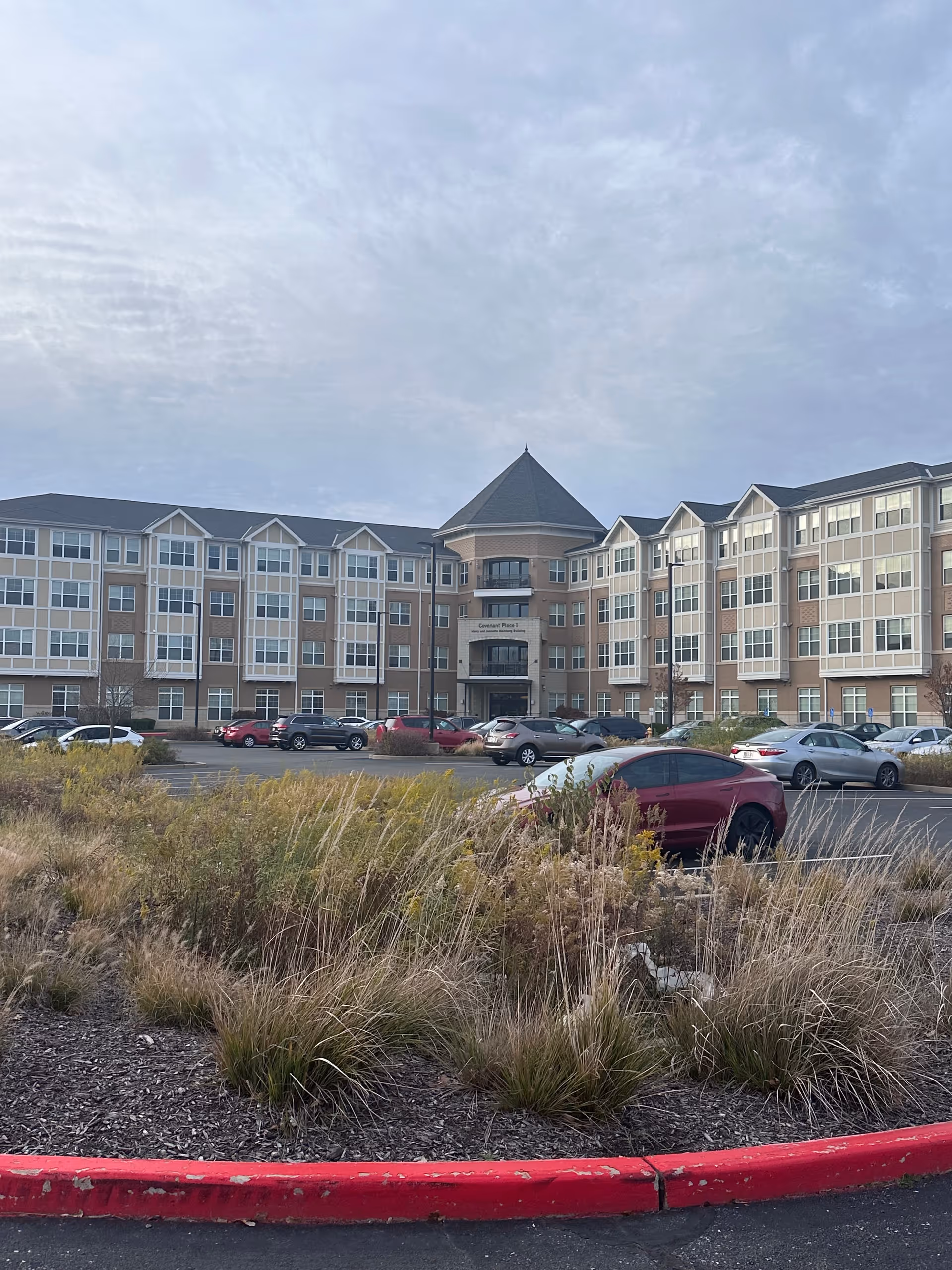 Exterior view of a large multi-story senior living facility building with a peaked roof section in the center. There is a parking lot in front with several cars parked and landscaped areas with tall grasses and plants near the curb.