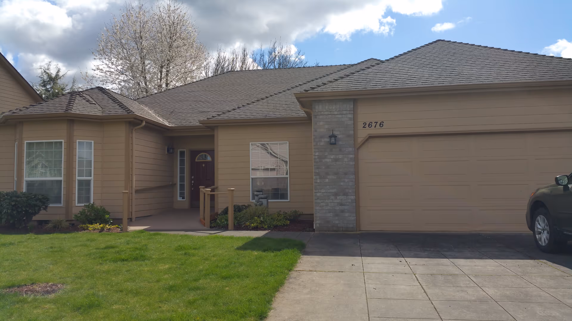 Front exterior view of a single-story house with beige siding, a two-car garage, a small porch with a ramp, and a well-maintained green lawn under a partly cloudy sky.
