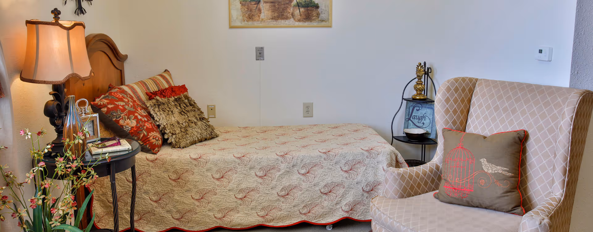A cozy bedroom featuring a single bed with a patterned quilt and multiple decorative pillows. Next to the bed is a small round side table with a lamp, picture frame, and other small items. A comfortable armchair with a decorative pillow is positioned nearby. The walls are light-colored with a framed picture hanging above the bed.