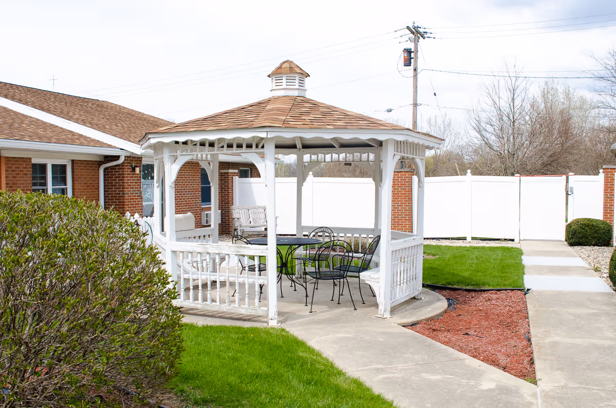 Outdoor gazebo with a wooden shingle roof and white railings, containing a black metal table and chairs, situated on a concrete patio surrounded by green grass, bushes, and a white fence, with a brick building in the background.