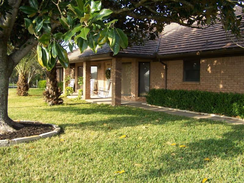 Exterior view of a single-story brick building with a covered porch featuring rocking chairs. The building is surrounded by a well-maintained lawn, bushes, and trees, including a palm tree and a tree with broad green leaves in the foreground.