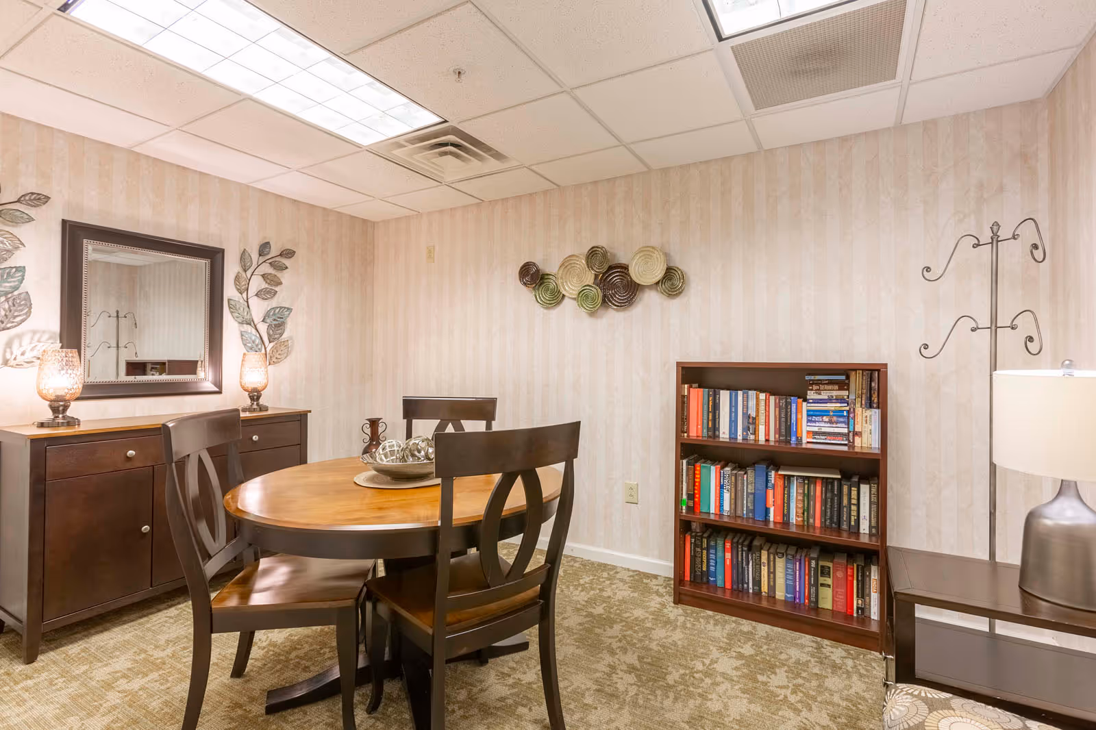 A cozy interior room with a round wooden table surrounded by four dark wooden chairs. Against the wall is a dark wooden sideboard with two decorative lamps and a mirror above it. A bookshelf filled with books is on the right side, next to a metal coat rack and a table lamp. The walls have a subtle striped wallpaper and the floor is carpeted.