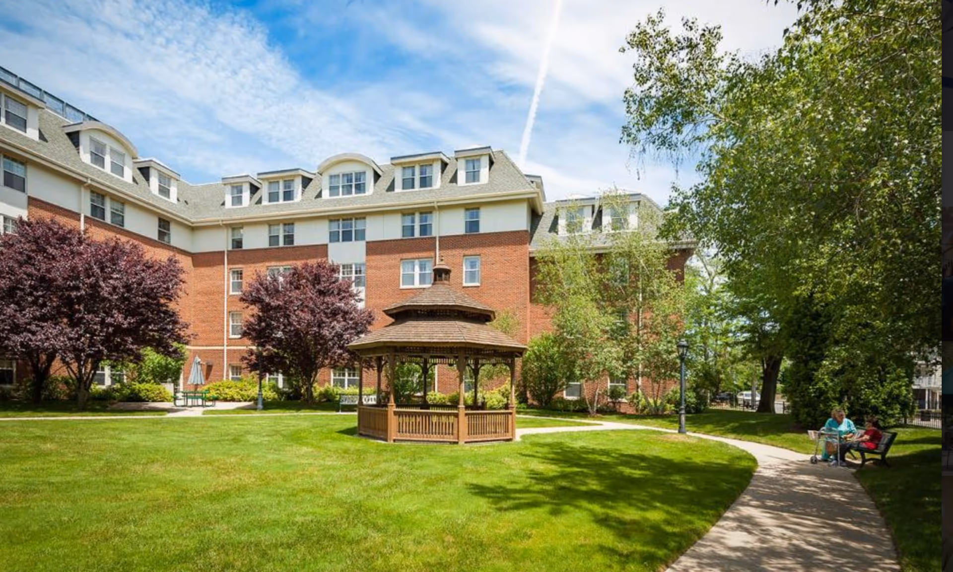 Outdoor view of Providence House Assisted Living showing a well-maintained lawn with a wooden gazebo in the center, surrounded by trees and a brick multi-story building in the background. Two people are sitting on a bench along a curved pathway on the right side.