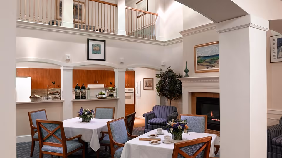 A cozy common area in a senior living facility featuring two tables with white tablecloths and floral centerpieces, surrounded by wooden chairs with blue cushions. In the background, there is a kitchen area with wooden cabinets and a white refrigerator. To the right, a fireplace with a painting above it and a striped armchair next to a potted plant are visible. The room has high ceilings with a balcony railing above.