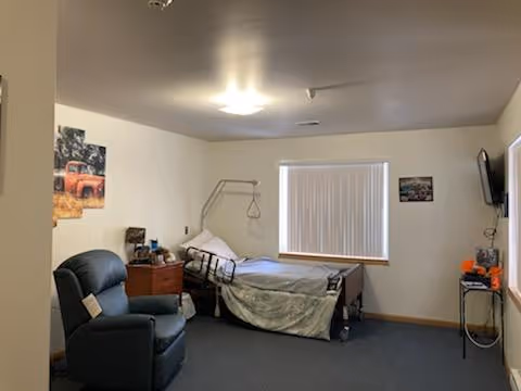 A simple, clean bedroom in a senior living facility with a hospital-style bed, a recliner chair, a small wooden nightstand, a window with closed blinds, a wall-mounted TV, and a few framed pictures on the walls.