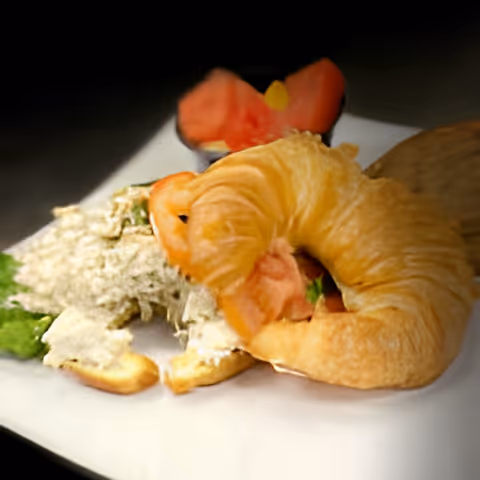 Close-up of a plate with a croissant sandwich filled with lettuce and tomato, accompanied by a small serving of a creamy salad and a slice of tomato in the background.