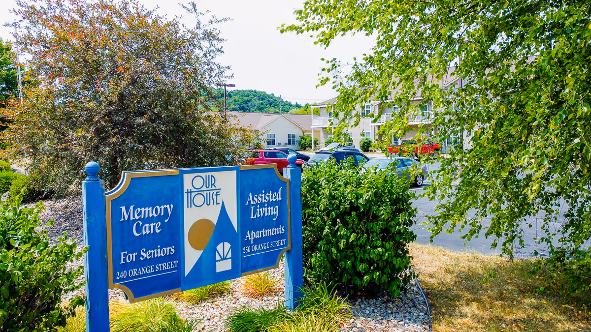 Outdoor view of a blue and white sign for Our House Senior Living showing Memory Care for Seniors at 240 Orange Street and Assisted Living Apartments at 250 Orange Street, surrounded by greenery and bushes with a parking lot and senior living buildings in the background.