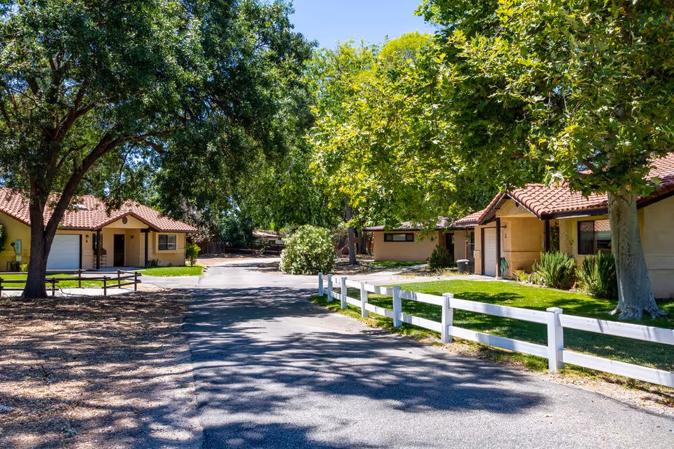 A sunny residential street within Oakview Village Independent Senior Living, featuring single-story houses with red tile roofs, surrounded by green trees and lawns, and a white wooden fence along the sidewalk.