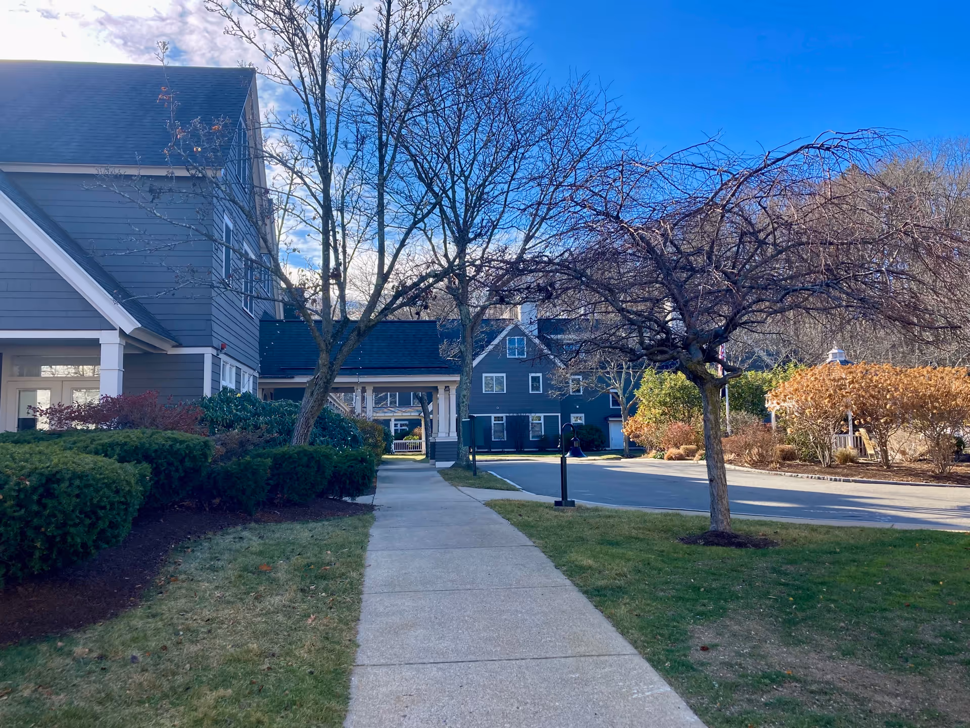 Sidewalk leading through landscaped grounds with leafless trees and bushes, adjacent to a gray multi-story building under a clear blue sky.
