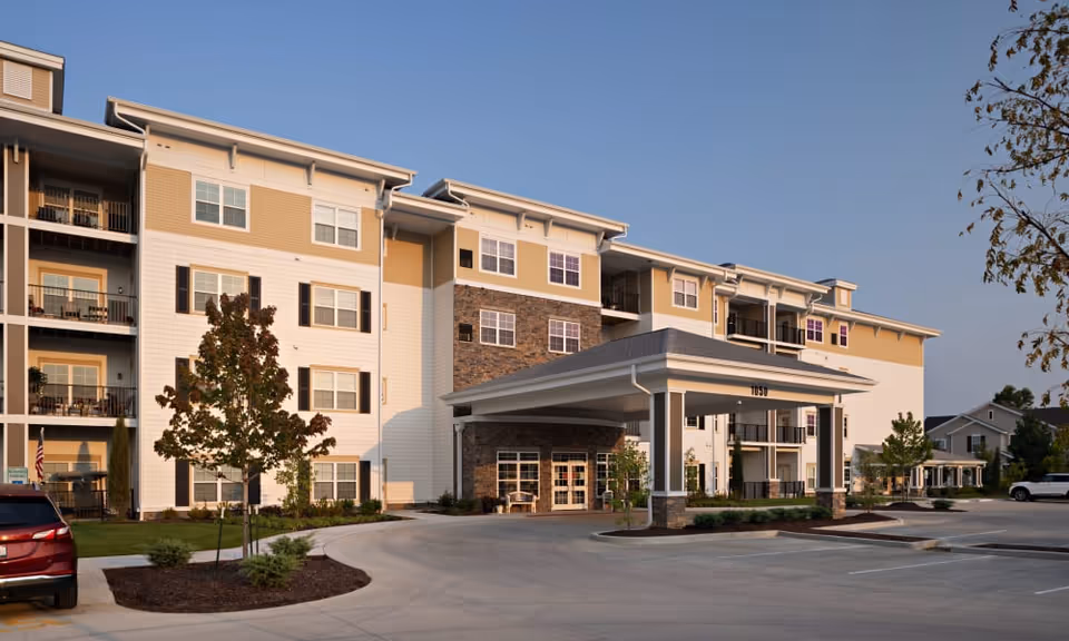 Exterior view of a multi-story senior living facility building with beige and white siding, stone accents, balconies, and a covered entrance driveway. There are a few small trees and landscaped areas around the parking lot, and a clear blue sky overhead.