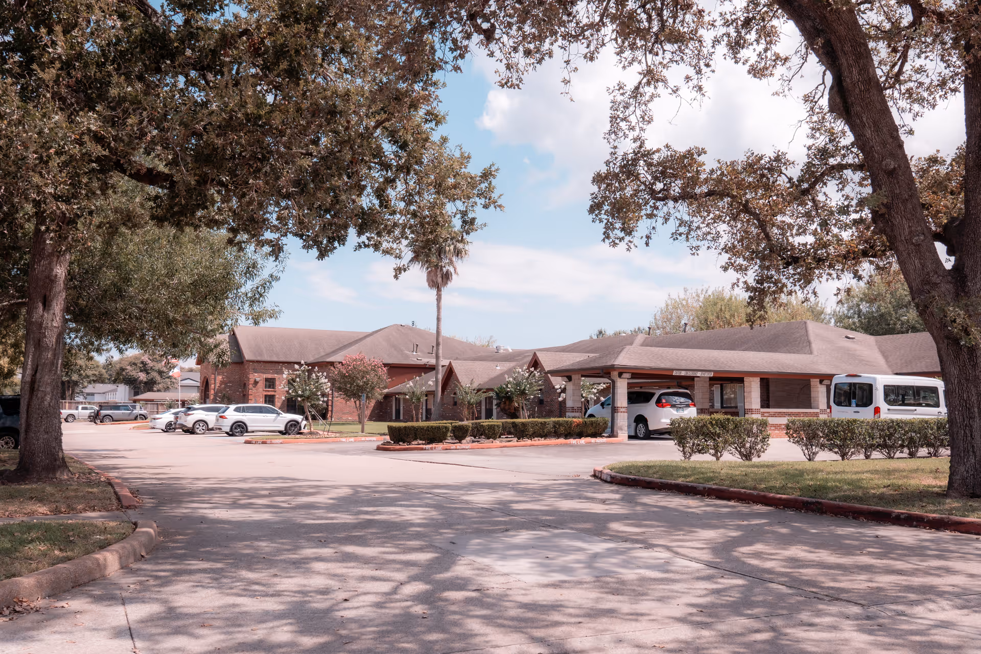 Exterior view of a single-story brick building with a covered entrance and several parked cars in front. The scene includes large trees with branches extending overhead, casting shadows on the driveway and parking area. The sky is partly cloudy.
