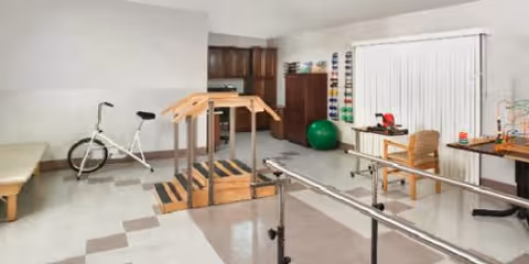 A physical therapy or rehabilitation room with exercise equipment including a stationary bike, parallel bars, a wooden staircase for step exercises, an exercise ball, and various therapy tools on shelves and tables. The room has white walls, a window with vertical blinds, and wooden cabinetry.