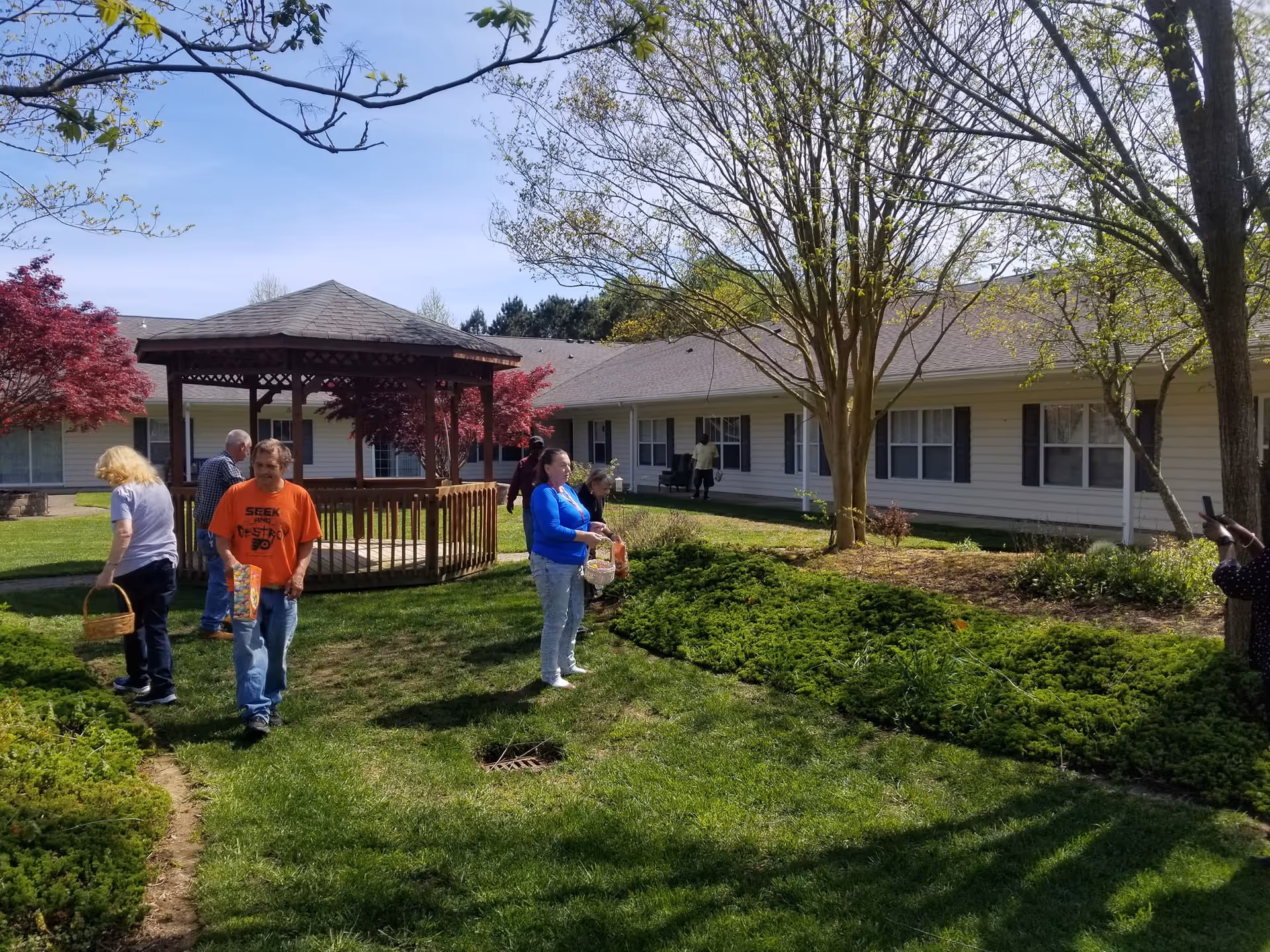 A group of people outdoors in a grassy courtyard area with a wooden gazebo. The courtyard is surrounded by a single-story building with white siding and multiple windows. Trees with budding leaves and red foliage bushes are visible, and the sky is clear and blue.