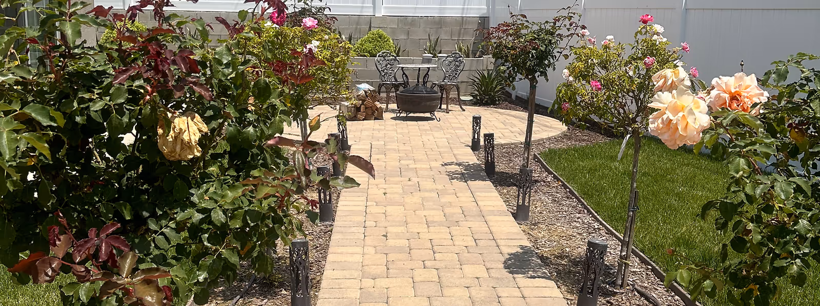 A paved outdoor walkway lined with blooming rose bushes on both sides leading to a small seating area with metal chairs and a fire pit. The area is surrounded by a white fence and a well-maintained lawn.