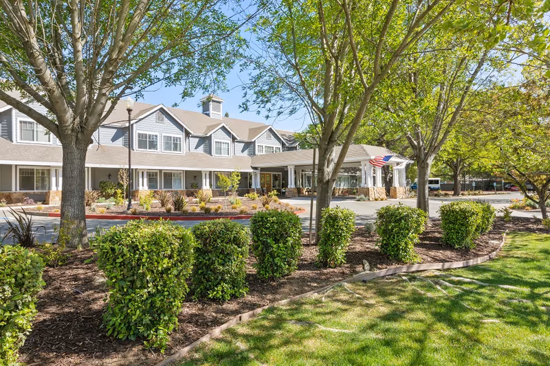 Exterior view of a senior living facility building with a covered entrance, surrounded by trees, bushes, and a well-maintained lawn under a clear blue sky.