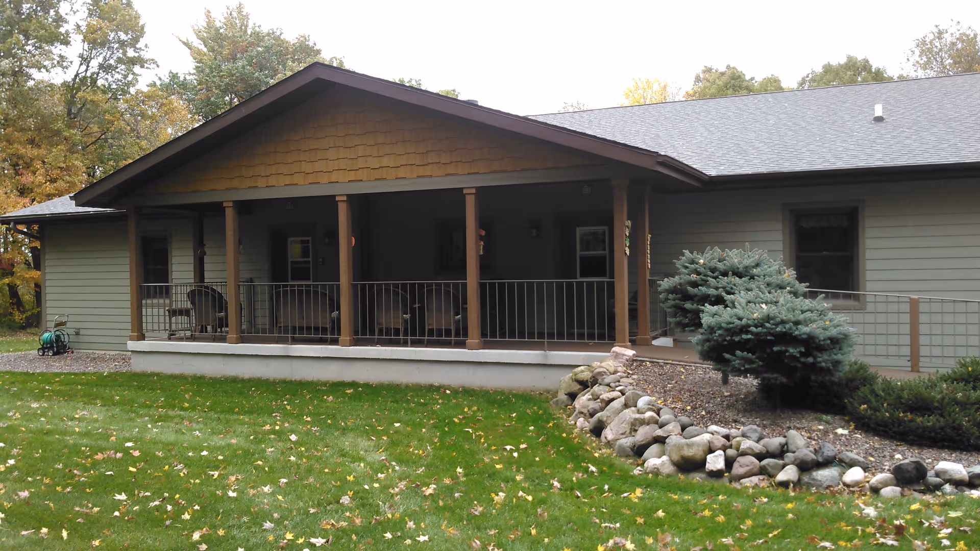 Exterior view of a single-story building with a covered porch supported by wooden columns. The porch has outdoor seating and is surrounded by a metal railing. The building is surrounded by green grass, a rock garden with small shrubs, and trees with autumn foliage in the background.