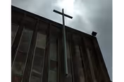 A tall metal cross mounted on the roof of a building with vertical windows, under a cloudy sky.