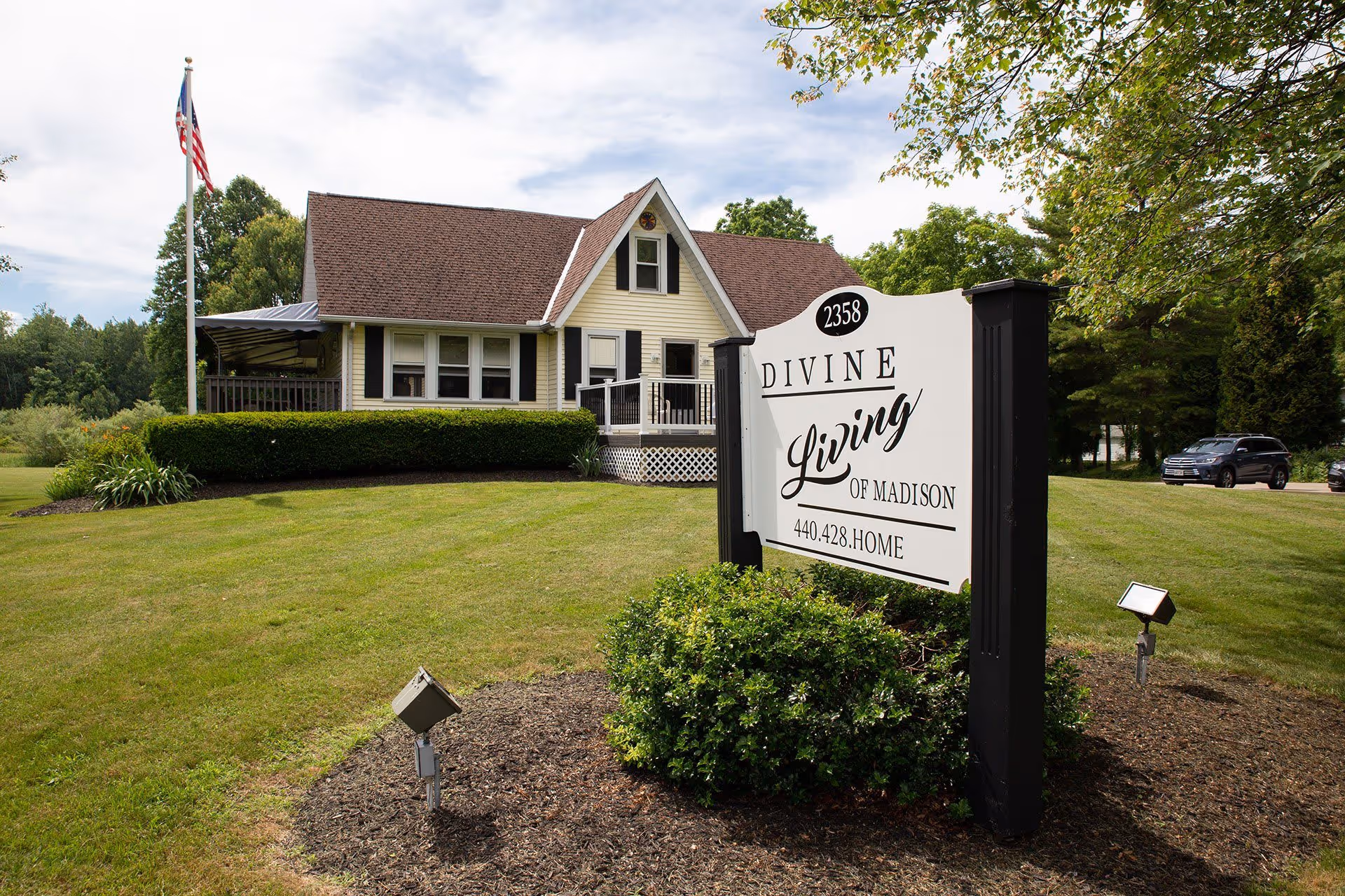 Exterior view of a single-story house with beige siding and a brown roof, surrounded by a well-maintained lawn and bushes. In the foreground, there is a white sign with black posts that reads '2358 Divine Living of Madison 440.428.HOME'. An American flag on a flagpole is visible to the left of the house, and a few cars are parked on the right side in the background.