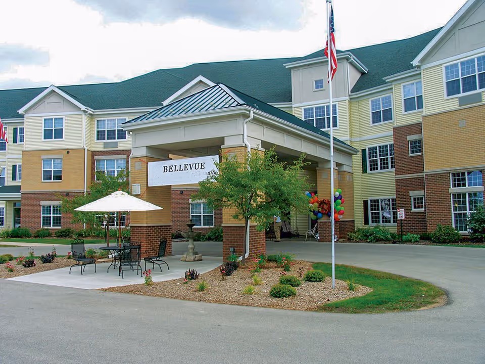 Exterior view of Bellevue Senior Living facility showing a three-story building with a covered entrance. There is a small patio area with a table, chairs, and an umbrella, surrounded by landscaped plants and a flagpole with an American flag.