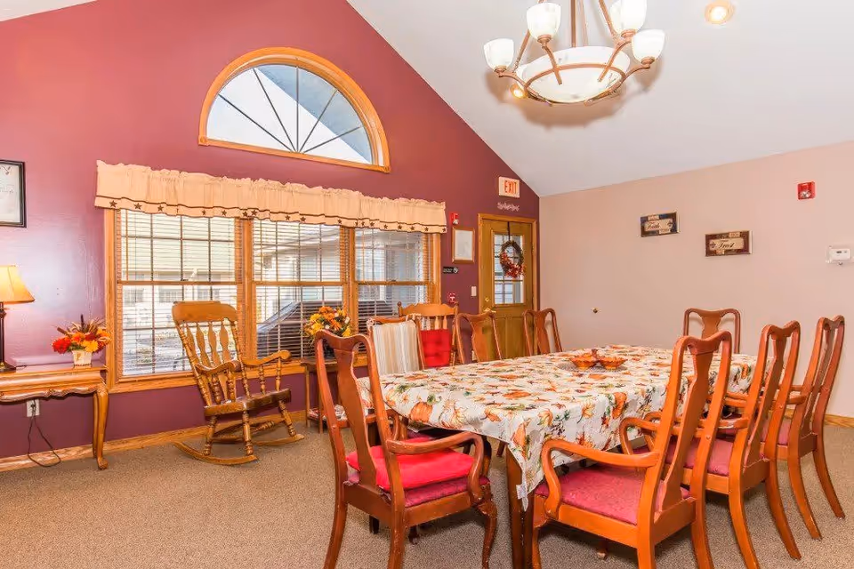 A dining room with a long wooden table covered with a floral tablecloth. The table is surrounded by wooden chairs with red cushions. There is a wooden rocking chair near a window with wooden blinds and a valance. The walls are painted in two colors: a deep maroon on one side and beige on the other. A chandelier with multiple lights hangs from the ceiling. There are decorative items including a small lamp on a side table, flower arrangements, and framed wall art. An exit door with a wreath is visible in the background.