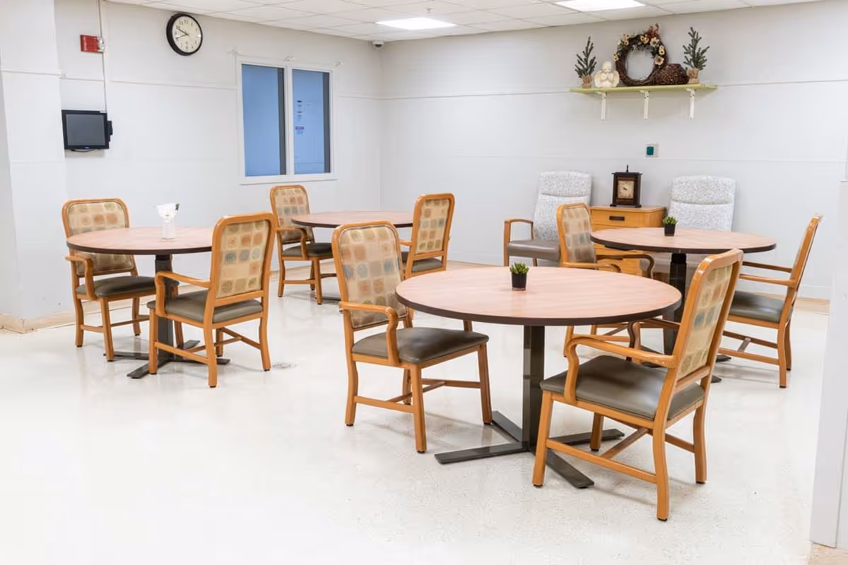 A clean and bright dining area with four round wooden tables, each surrounded by four wooden chairs with cushioned seats and backs. The room has white walls and a white floor, a clock on the wall, a small window, and a shelf with decorative items including a wreath and a small basket. Two upholstered armchairs and a small wooden cabinet are visible in the background.