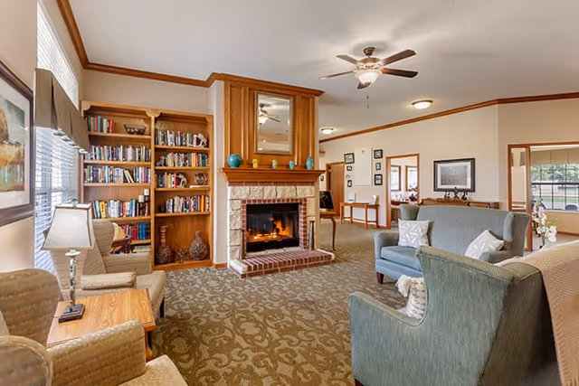 A cozy living room area in a senior living facility featuring a lit brick fireplace with a wooden mantle and mirror above it. There are built-in wooden bookshelves filled with books and decorative items to the left of the fireplace. The room has patterned carpet, multiple upholstered chairs and sofas with cushions, a wooden side table with a lamp, and ceiling fans with lights. Large windows allow natural light to brighten the space.