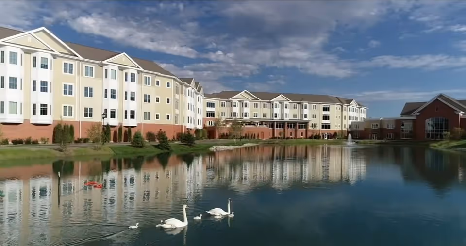 A large multi-story senior living facility named The Moorings is shown with a pond in the foreground. The building has a beige and white exterior with multiple windows and a brick base. The pond reflects the building and sky, and there are swans and ducks swimming on the water. The sky is partly cloudy with blue patches.