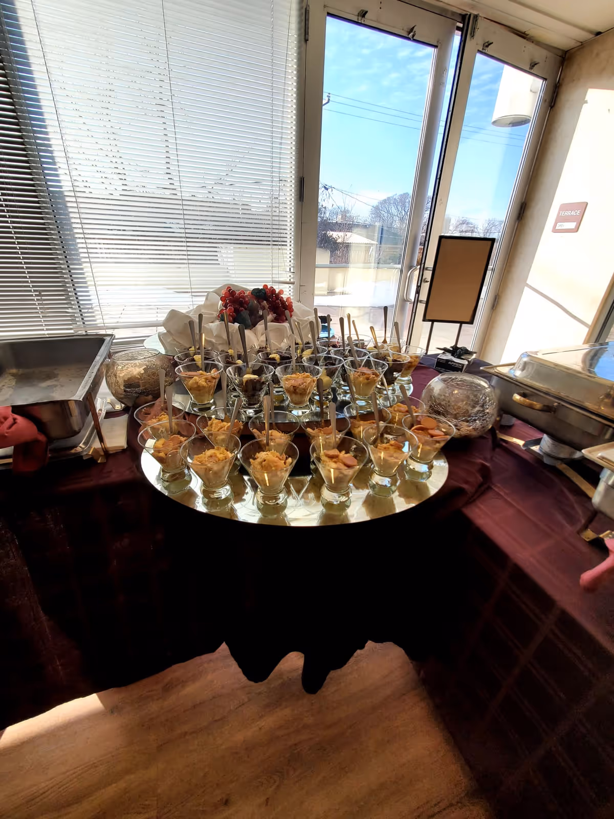 A buffet table set up with multiple small glass cups filled with a dessert or appetizer, each with a spoon. The table is covered with a dark tablecloth and is positioned near a window with blinds. Outside the window, a bright sunny day is visible. There are also chafing dishes on either side of the table and a small sign on a stand.
