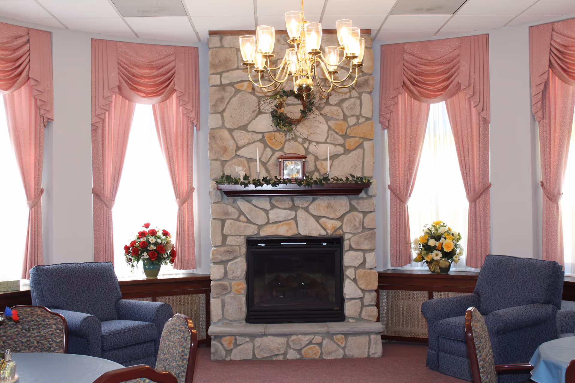 A cozy living room area with a stone fireplace in the center, decorated with a wreath and candles on the mantel. There are three tall windows with pink drapes and valances, and flower arrangements on the window sills. Two blue upholstered armchairs are positioned on either side of the fireplace, with a round table and chairs partially visible in the foreground. A brass chandelier hangs from the ceiling above.