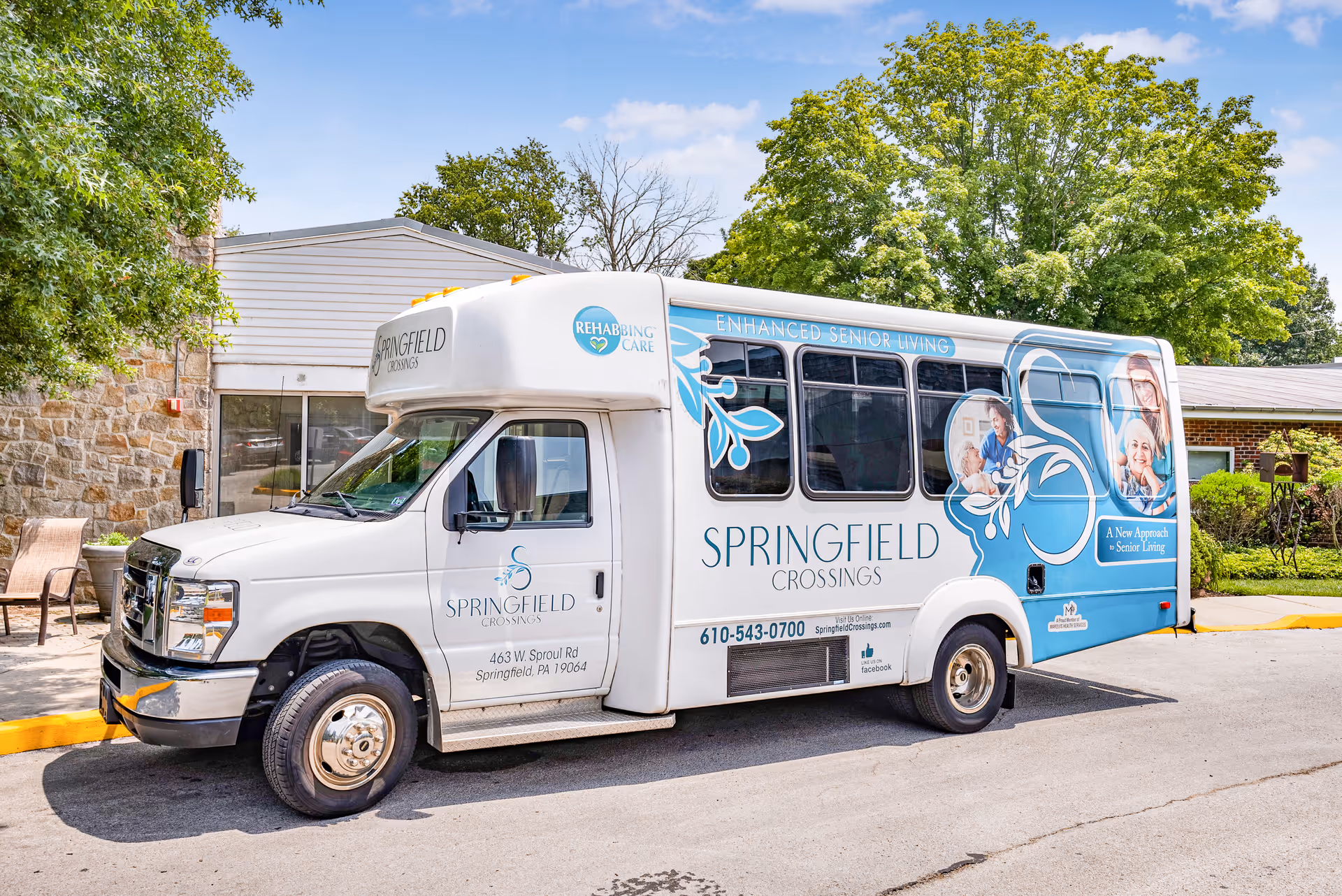 A white and blue shuttle bus parked outside a building with trees and greenery in the background. The bus is branded with Springfield Crossings logos and text promoting enhanced senior living and rehab care. The bus also displays contact information and images of seniors and caregivers.