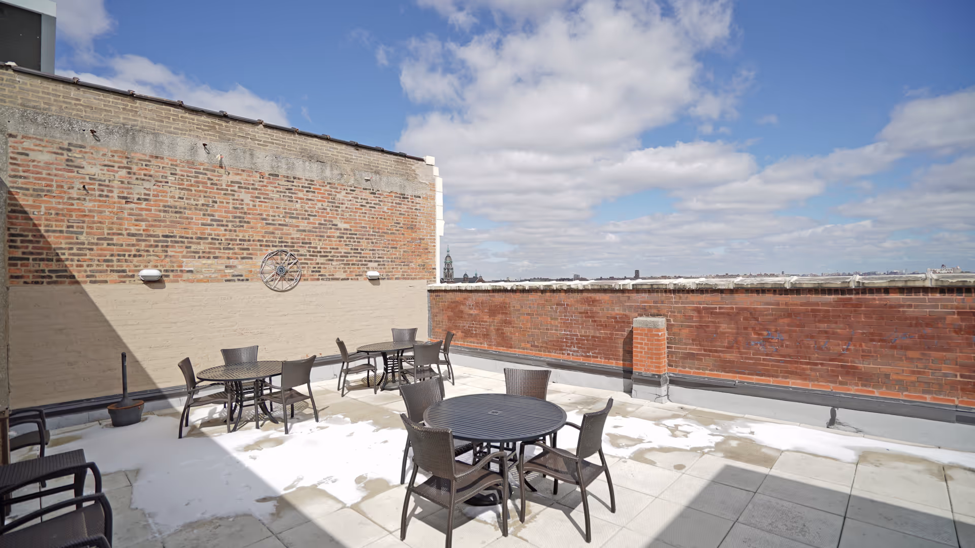 Outdoor rooftop patio area with several round black metal tables and matching chairs. The patio is surrounded by brick walls and has a clear blue sky with some clouds overhead.