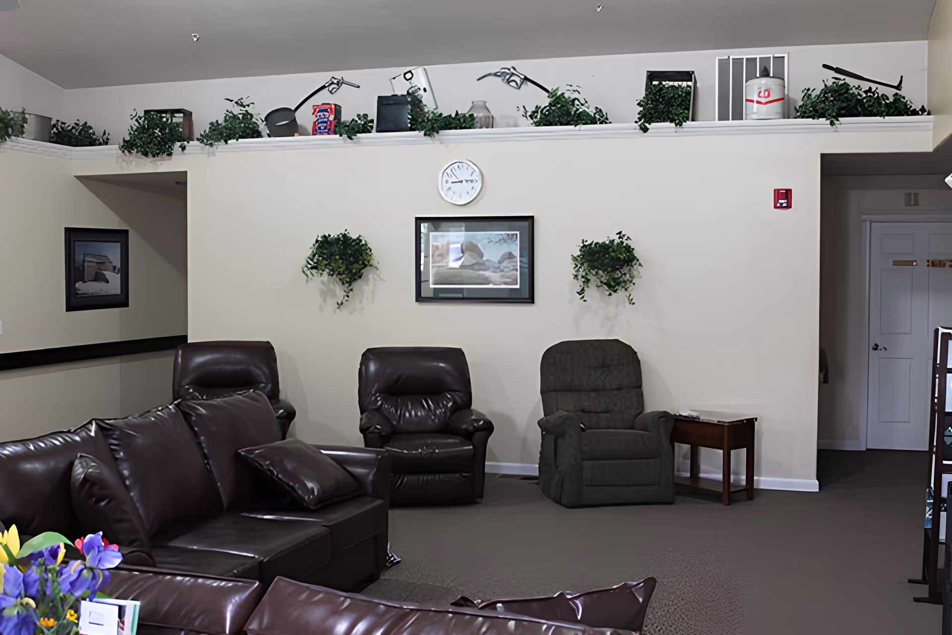 A cozy living room area with dark brown leather sofas and two recliner chairs, one brown and one gray. The walls are light-colored and decorated with two hanging green plants and a framed picture beneath a round wall clock. Above the wall is a ledge with various decorative items and more greenery. There is a small wooden side table next to the gray recliner and a hallway entrance visible on the right side.