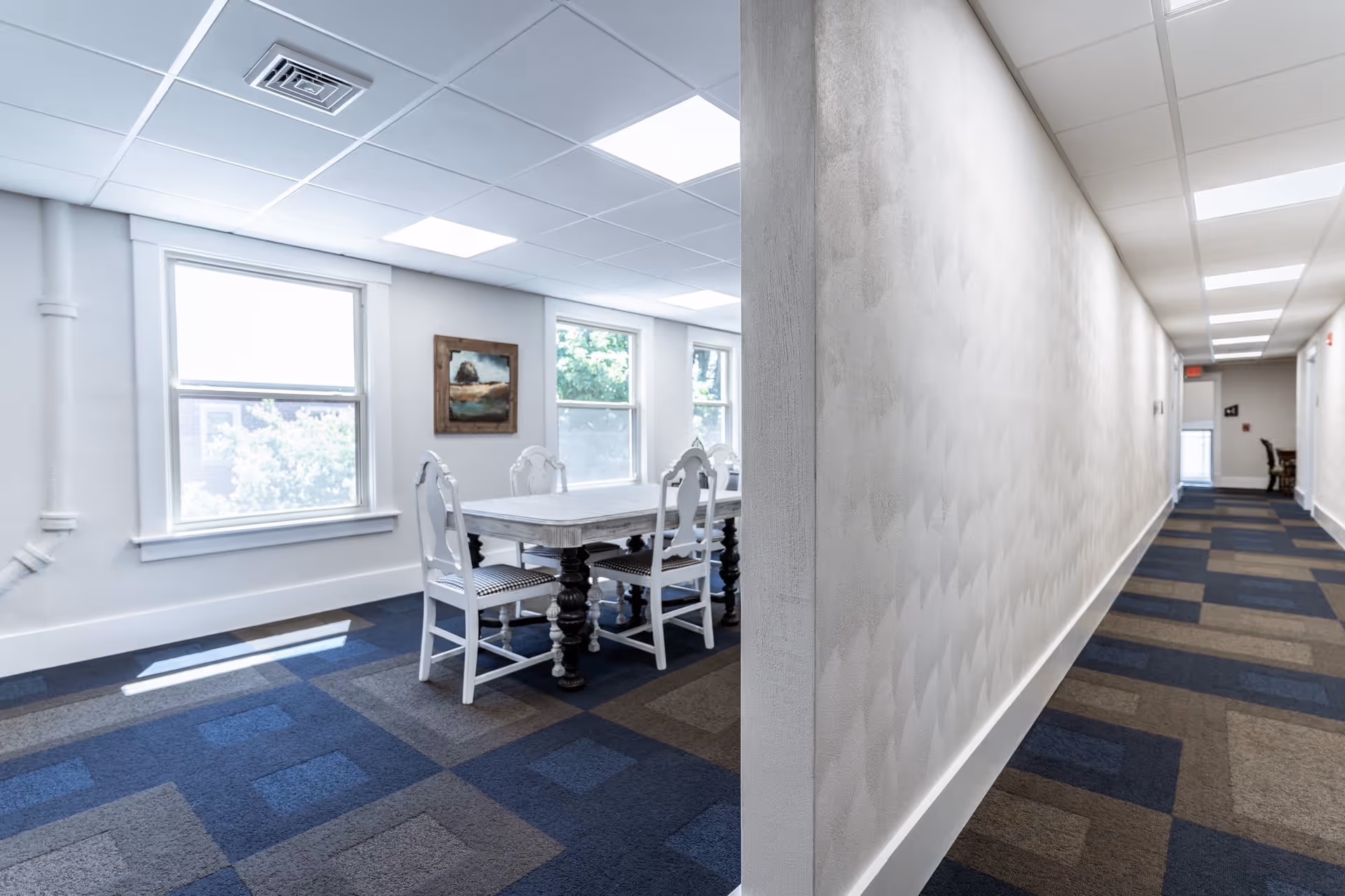 A bright interior space featuring a hallway with patterned carpet leading to a small dining area with a white wooden table and four matching chairs. The dining area has three large windows letting in natural light and a framed painting on the wall.