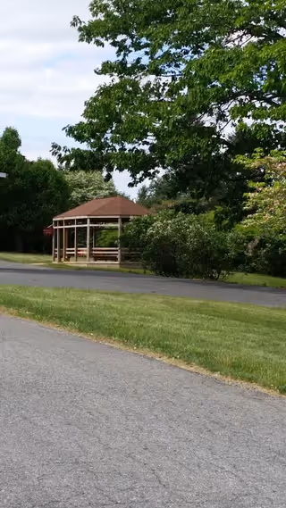 A wooden gazebo with a brown roof situated in a green outdoor area with trees and bushes surrounding it. There is a paved road and a grassy lawn in the foreground under a partly cloudy sky.