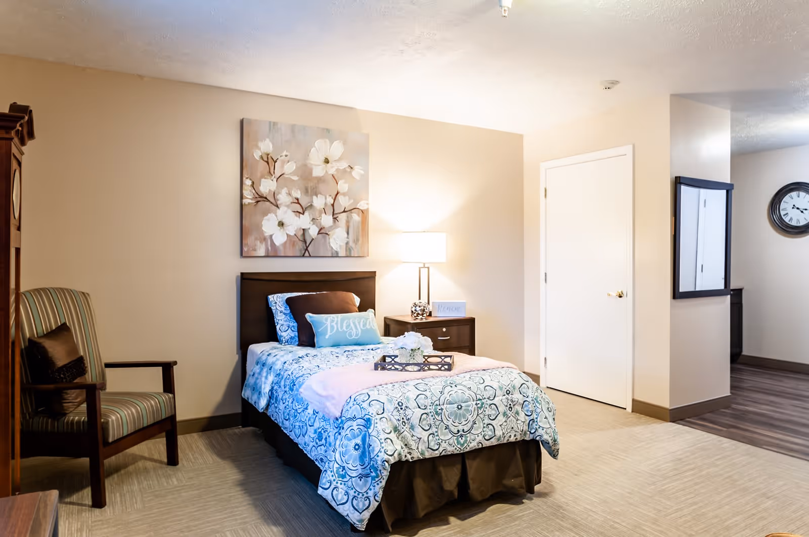 A cozy bedroom in a senior living facility featuring a single bed with a blue and white patterned comforter, decorative pillows including one that says 'Blessed', a wooden nightstand with a lamp and a small sign, a striped armchair with a dark cushion, a large floral painting above the bed, and a wall clock visible in the adjacent hallway.