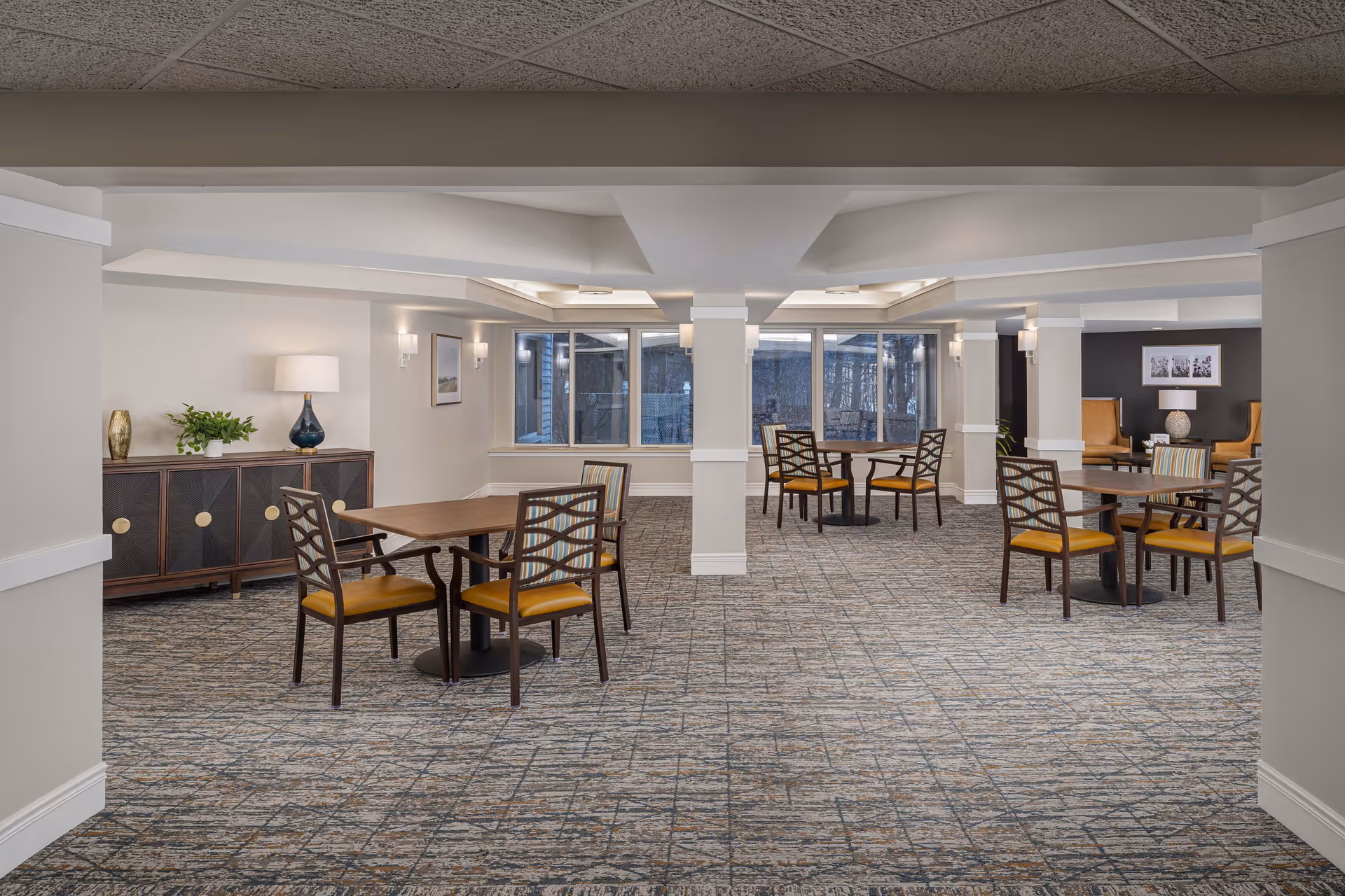 A spacious senior living common area with multiple wooden tables and chairs arranged on a patterned carpet. The room features white walls, large windows letting in natural light, and decorative lamps on side tables and a cabinet. There are columns dividing the space and comfortable seating areas in the background.