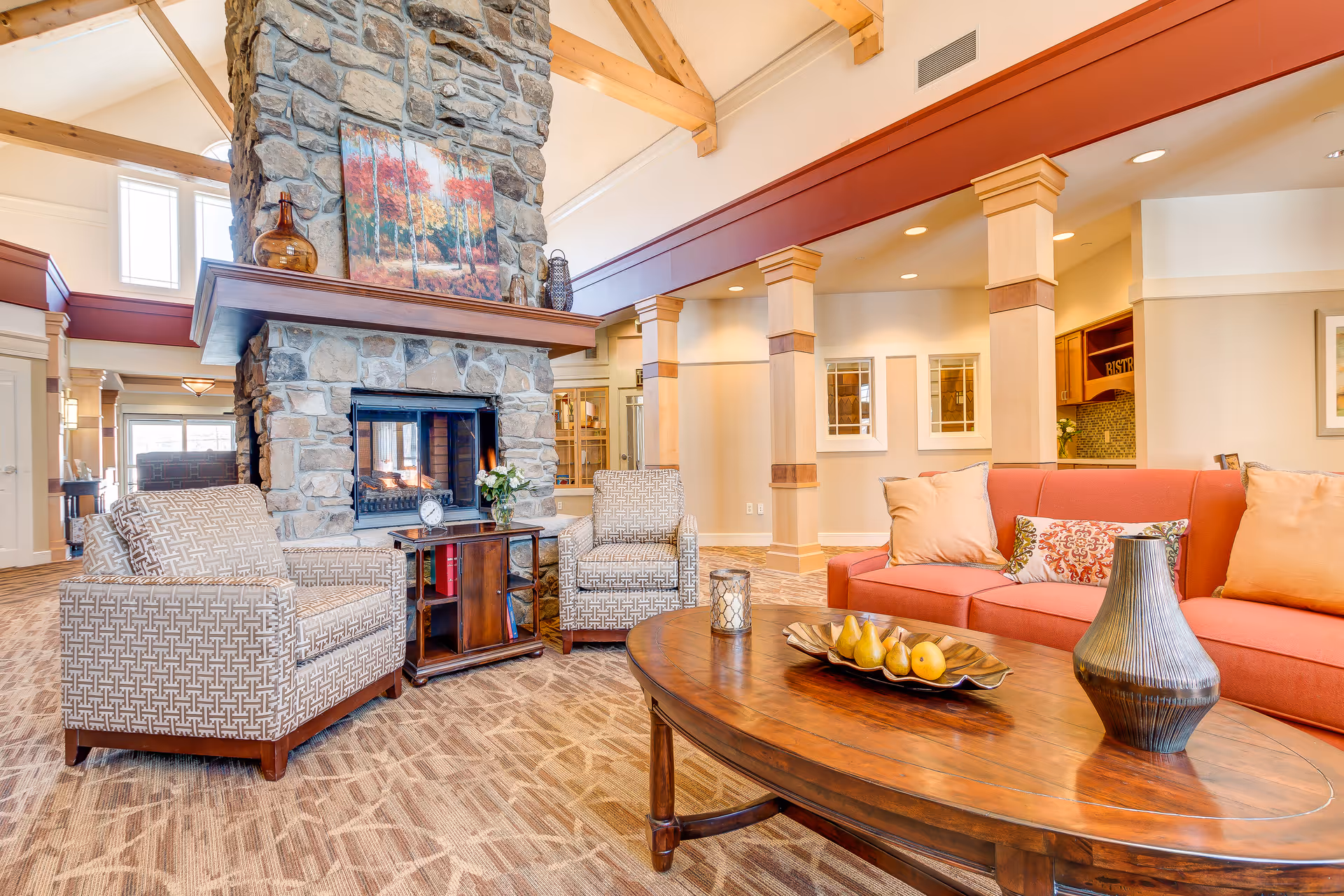 A cozy living room area in a senior living facility featuring a stone fireplace with a colorful autumn forest painting above it. The room has two patterned armchairs and a coral-colored sofa with multiple cushions. A wooden coffee table in front of the sofa holds a decorative vase, a candle, and a tray with pears. The space is well-lit with natural light from large windows and has wooden beams on the ceiling.