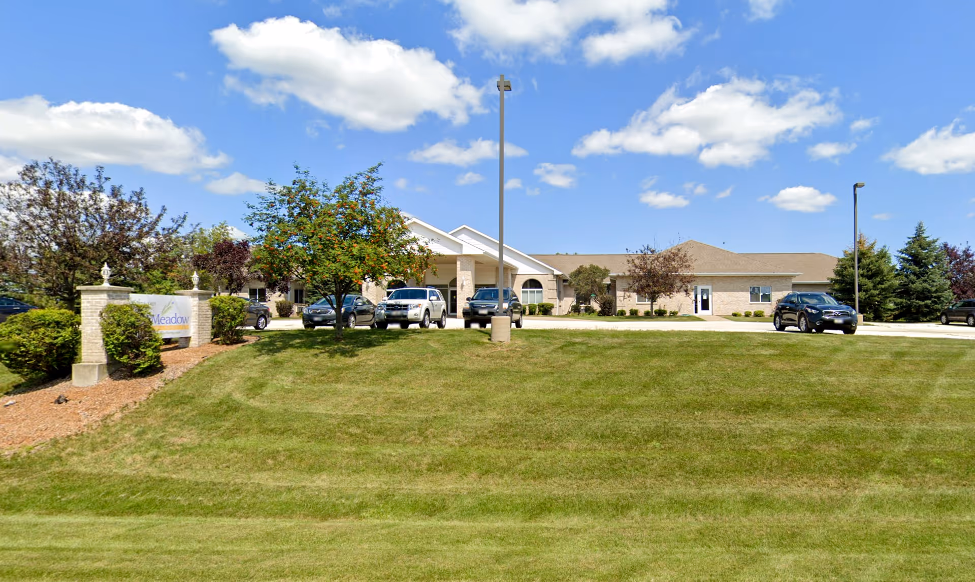 Exterior view of Sage Meadow De Pere facility showing a single-story building with a beige brick facade, several parked cars, a green lawn, trees, and a blue sky with scattered clouds.