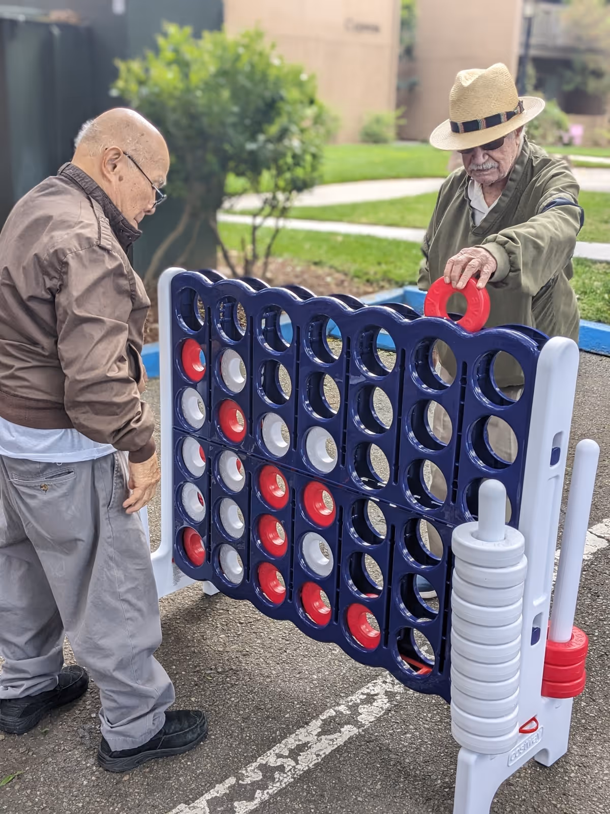 Two elderly men playing a large outdoor Connect Four game on a paved area with grass and bushes in the background. One man is placing a red game piece into the game while the other watches.