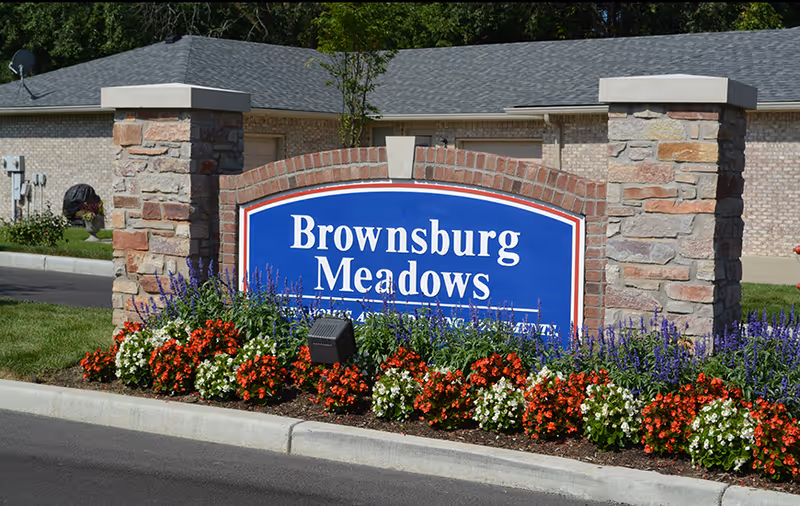 A large blue sign with white text reading 'Brownsburg Meadows' set between two stone pillars. The sign is surrounded by a flower bed with red, white, and purple flowers. In the background, there is a brick building with a gray roof.