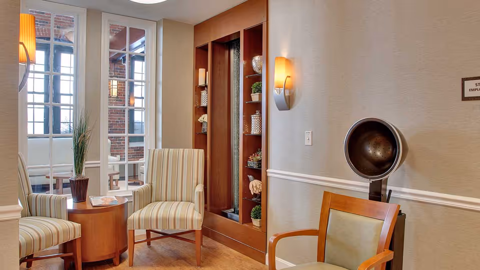 A cozy sitting area with two striped armchairs and a round wooden table between them, holding a potted plant and a magazine. Behind the chairs is a large window with multiple panes, showing a brick wall outside. To the right, there is a vintage hair dryer chair and a built-in wooden shelf with decorative items. The walls are light-colored with a white chair rail and a wall sconce providing warm lighting.